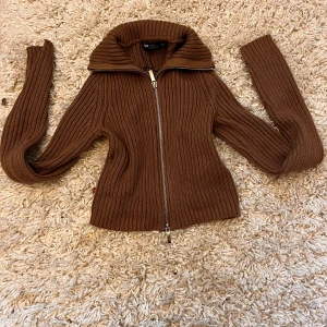 Brun stickad kofta med dragkedja - Säljer en snygg brun stickad kofta med dragkedja framtill. Den har en hög krage och långa ärmar, perfekt för höst och vinter. Koftan är ribbad vilket ger den en stilren look. Passar både till vardags och lite finare tillfällen.