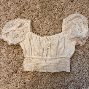 Beige blus med puffärmar - Säljer en superfin vit blus med puffiga kortärmar och broderade detaljer. Den har en söt knytning framtill och är croppad med en smockad midja. Perfekt för sommardagar eller en festlig kväll!