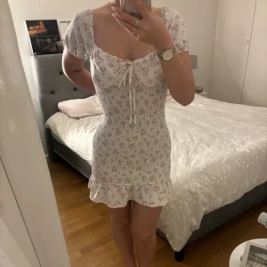 Blommig kortklänning från Shein - Säljer en superfin blommig kortklänning från Shein. Den är vit med små blommor i rosa och grönt. Klänningen har korta puffärmar och en söt knytning framtill. Perfekt för sommardagar eller en mysig vårutflykt! 🌸