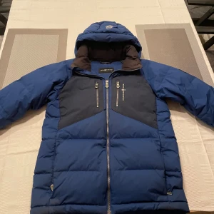 Sail racing jacka - Säljer en snygg blå pufferjacka från Sail Racing. Jackan har en cool design med mörkare paneler på bröstet och två dragkedjefickor framtill. Den är perfekt för kalla vinterdagar och har en skön huva för extra värme. Jackan är i bra skick.