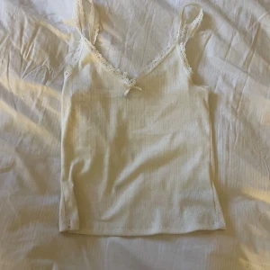 Vit linne från H&M Divided - Säljer ett supergulligt vitt linne från H&M Divided. Det är i storlek XS och har en söt spetsdetalj vid halsen samt en liten rosett. Materialet är ribbat och känns mjukt och skönt mot huden. Perfekt för sommardagar eller som ett lager under en tröja.