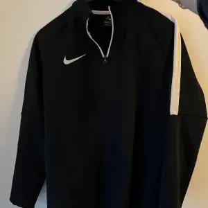 Säljer en svart tröja från Nike med vit dragkedja och vita detaljer på ärmarna. Perfekt för träning eller vardagsbruk. Tröjan har långa ärmar och en sportig look. Använd ett par gånger och är i bra skick. Tröjan är märkt som för 12-13 åringar.