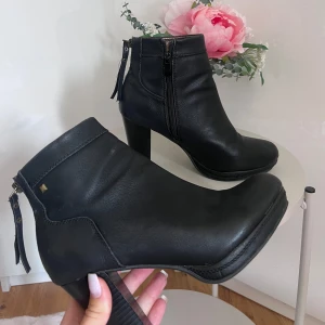Svarta stövletter med klack och dragkedja - Säljer ett par snygga svarta stövletter i skinn med hög klack. De har en praktisk dragkedja på sidan och en dekorativ dragkedja baktill. Perfekta för höst och vinter, och funkar lika bra till vardags som till fest. De är i bra skick och redo för en ny ägare!
