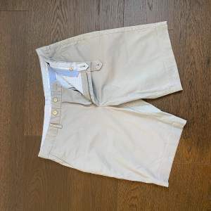 Säljer ett par stilrena beige shorts i bra skick. De har en klassisk design med knappstängning och bälteshällor. Perfekta för sommardagar eller en avslappnad vårlook. Shortsen har också två bakfickor med knappstängning för extra stil och funktionalitet.