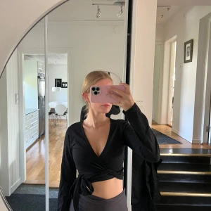 Svart omlottopp - Säljer en snygg svart omlottopp med långa ärmar. Toppen har en djup v-ringning och knyts i midjan för en tight passform. Perfekt för både vardag och fest!