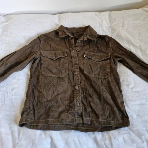 Brun denimskjorta från Levi's - Säljer en snygg brun denimskjorta från Levi's i storlek small (passar som en XS). Skjortan är tillverkad av 100% bomull och har en cool, lite sliten look. Den har långa ärmar och två bröstfickor med knappar, ett väldigt blekt tropiskt print. Perfekt för en avslappnad stil!