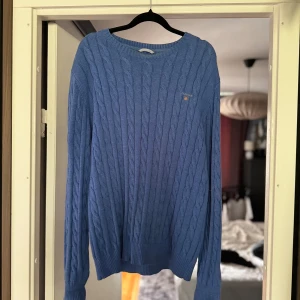 Blå stickad tröja från GANT - Säljer en snygg blå stickad tröja från GANT. Den har ett klassiskt flätmönster och en liten broderad logga på bröstet. Perfekt för kyliga dagar och passar både till vardags och lite finare tillfällen. Tröjan är i storlek M och är i mycket bra skick.