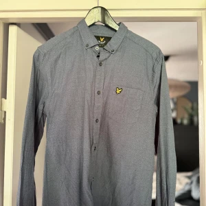 Blå skjorta från Lyle & Scott - Säljer en snygg blå skjorta från Lyle & Scott. Den har en klassisk krage och långa ärmar, perfekt för både vardag och fest. Skjortan är dekorerad med det ikoniska gula logotypen på bröstet och har knappar hela vägen ner. Materialet känns bekvämt och stilrent.