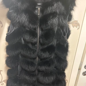 Svart pälsväst med dragkedja - Säljer en superfin svart pälsväst som är perfekt för att hålla sig varm och stilren. Västen har en dragkedja framtill och är i ett mjukt och fluffigt material. Den är perfekt att bära över en tröja för extra värme och stil.