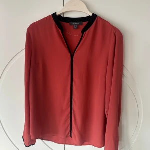 Röd blus från Primark - Säljer en snygg röd/rosa(rost) blus från Primark i storlek 42. Den har en stilren design med svarta detaljer längs kragen och knäppningen. Perfekt för både vardag och fest! 💃