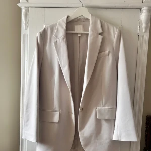 Vit kavaj från H&M - Säljer en stilren vit kavaj från H&M i storlek XS. Kavajen har en klassisk design med knappar framtill och fickor. Perfekt för både vardag och festliga tillfällen. Passar bra till både jeans och klänning!