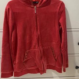 Röd hoodie med matchande byxor - Säljer en supermjuk och bekväm pyjamas. Överdelen har långa ärmar och praktiska fickor framtill. Perfekt för en avslappnad dag hemma. Byxorna har en elastisk midja och dekorativa detaljer. Använd, köpt för 400kr