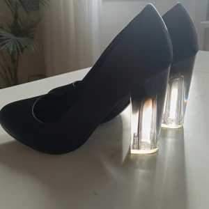 Svarta pumps med transparent klack - Säljer ett par eleganta svarta pumps med en unik transparent klack. Skorna är i nyskick och har en stilren design som passar perfekt för festliga tillfällen. Materialet är mjukt och bekvämt, vilket gör dem sköna att bära hela kvällen. 