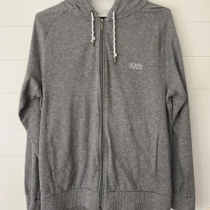 Grå hoodie från Hugo Boss - Säljer en stilren grå hoodie från Hugo Boss. Den har en dragkedja framtill och en justerbar huva med snören, snörena har tyvärr börjat snurra upp sig.  Tröjan är välanvänd men i övrigt i gott skick