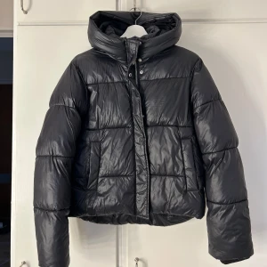 Glansig dunjacka - Glansig dunjacka från Nelly i storlek 36. Liten defekt se bild 3 samt 4 (den man hänger upp jackan på har lossnat). Annars jättefint skick men stor luva och hög i halsen. Skriv vid fler bilder💗