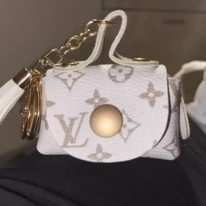 Vit mini handväska med monogram - Säljer en superfin vit mini handväska med monogrammönster. Den har en snygg guldfärgad kedja och en liten tofsdetalj. Perfekt som en liten accessoar eller nyckelring. Väskan har en tryckknapp för att hålla den stängd.
