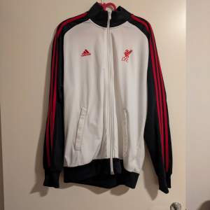 Säljer en snygg vit och svart Adidas jacka med Liverpool FC-logga på bröstet. Jackan har röda detaljer och klassiska Adidas-ränder längs ärmarna. Den är långärmad och har en dragkedja framtill. Perfekt för fotbollsfans som vill visa sitt stöd för laget!