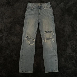 Blå jeans med slitningar från Perfect Jeans - Säljer ett par snygga blå jeans från Gina Tricot Perfect Jeans i storlek 38. De har en cool, sliten look med hål på knäna. Jeansen är i en klassisk straight fit, är högmidjade och har en knappgylf. Perfekta för en casual outfit.