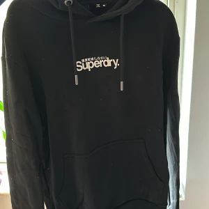 Svart hoodie från Superdry - Säljer en svart hoodie från Superdry i storlek M. Den har en klassisk design med Superdry-loggan tryckt på framsidan och justerbara snören i huvan. Perfekt för en avslappnad stil!