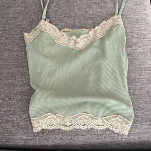 Grön ribbad linnetopp med spetsdetaljer - Säljer en superfin grön ribbad linnetopp med smala axelband. Toppen har vackra spetsdetaljer längs halsringningen och nederkanten, vilket ger den en romantisk touch. Perfekt för sommardagar eller som ett lager under en kofta. Aldrig använd!
