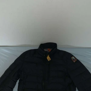 Svart dunjacka från Parajumpers - Säljer en snygg och varm svart dunjacka från Parajumpers. Jackan har en hög krage och en dragkedja framtill. Den är perfekt för kalla dagar och har en cool patch på ärmen. Passar både till vardags och lite finare tillfällen. Hör av dig om du är intresserad!