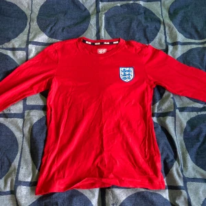 Röd England fotbollströja - Säljer en röd England fotbollströja med ett coolt emblem på bröstet. Tröjan är långärmad och perfekt för fotbollsfans. Den har bara vart använd ett få tal gånger och är i totalt ny skick 