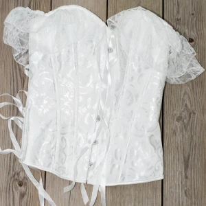 Vit korsett med spetsdetaljer - Säljer en elegant vit korsett med vackra spetsdetaljer och snörning baktill. Den har en romantisk känsla med sina offshoulder spetsärmar och ett subtilt blommönster. Perfekt för speciella tillfällen eller en festlig kväll. Helt ny i förpackning!