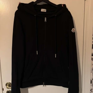Mörkblå hoodie från Moncler - Säljer en snygg svart hoodie från Moncler i nyskick. Den har en dragkedja framtill och en klassisk Moncler-logga på ärmen. Perfekt för höst och vår, med långa ärmar och en bekväm passform. Huvan har en stilren kant i rött och vitt. Passar både till vardags och lite finare tillfällen.