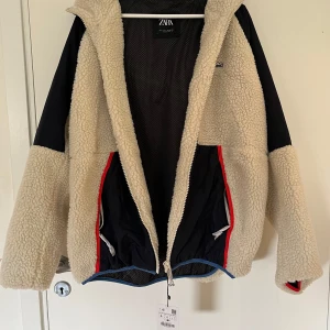 Teddy jacka från Zara - Den här jackan från Zara är en ny och oanvänd fleecejacka i storlek L. Jackan har en mjuk och varm fuskpäls, med en sportig design som inkluderar svarta inslag och röda detaljer längs med dragkedjan. Tyvärr blev den fel storlek, vilket är anledningen till att den säljs vidare.😇
