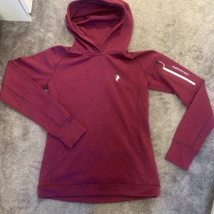 Vinröd hoodie från Peak Performance XS - Säljer en snygg vinröd hoodie från Peak Performance i storlek XS. Den har en praktisk dragkedjeficka på ärmen och är perfekt för kyliga dagar. Hoodien är i bra skick och superbekväm att ha på sig. Perfekt för både vardag och träning!