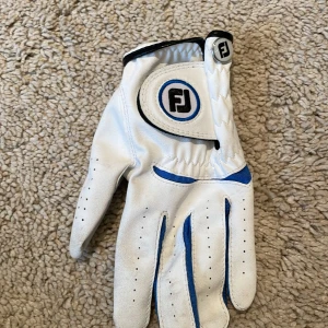Vit och blå handske från FootJoy - Säljer en vit handske från FootJoy med blå detaljer. Perfekt för golfare som vill ha bra grepp och stil. Handsken har en justerbar rem med FJ-logga och perforeringar för ventilation. Använd ett fåtal gånger, så den är i bra skick. Passar både för träning och tävling. 🏌️‍♂️