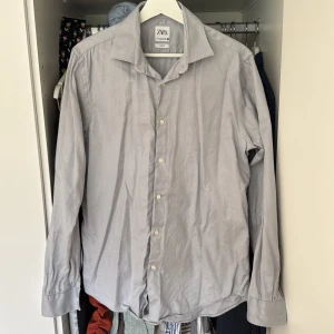 Grå slim fit skjorta från Zara - Säljer en stilren grå slim fit skjorta från Zara. Skjortan har långa ärmar och knappar framtill. Perfekt för både vardag och festliga tillfällen. Den är i bra skick och passar utmärkt till både jeans och kostymbyxor. Lättstrykning så ser den helt ny ut igen.