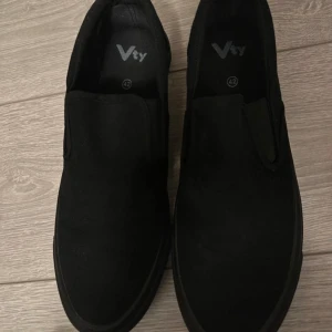 Svarta slip-on sneakers från Vty - Säljer ett par svarta slip-on sneakers från Vty i storlek 42. De är i nyskick och perfekta för både vardag och fest. Skorna har en enkel och stilren design utan snörning, vilket gör dem lätta att ta på och av. Passar bra till alla outfits!