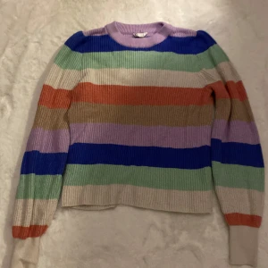 Flerfärgad randig stickad tröja - Säljer en färgglad stickad tröja med breda ränder i blått, grönt, lila, orange och beige. Tröjan har en rund hals och långa ärmar, perfekt för att liva upp garderoben. Den är mjuk och bekväm att bära.