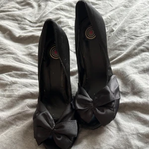 Svarta pumps med rosett - Eleganta svarta pumps i satin med en dekorativ rosett framtill. Skorna har en hög klack och en rundad tå, vilket ger en stilren look. Perfekta för en kväll ute eller en speciell tillställning. Köparen står för frakten