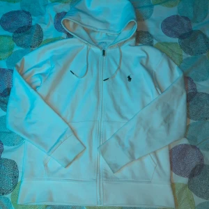 Vit huvtröja från Ralph Lauren - Säljer en stilren vit huvtröja från Ralph Lauren med dragkedja och det klassiska logotypbroderiet på bröstet. Tröjan har långa ärmar och en bekväm huva med dragsko. Perfekt för en avslappnad look.