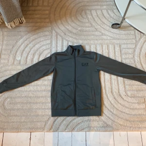 Grå jacka från Emporio Armani - Säljer en stilren grå jacka från Emporio Armani EA7. Jackan har en dragkedja framtill och långa ärmar. Perfekt för en casual look med sin enkla design och bekväma passform.