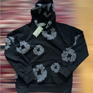 Svart hoodie från Denim Tears - Säljer en svart hoodie från Denim Tears med grå blommönster. Hoodien har en klassisk design med huva och långa ärmar. Tillverkad i USA och perfekt för en stilren look.
