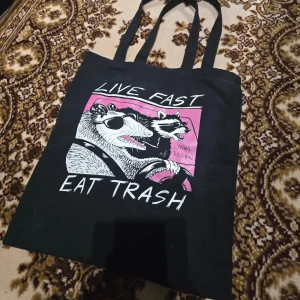 Totebag, tygväska m. Coolt tryck - Cool svart tygväsja med ett humoristiskt tryck av en opossum och en tvättbjörn i en bil. Texten 'Live Fast Eat Trash' ger en rolig och avslappnad känsla. Perfekt för att bära med sig vardagliga saker med stil.