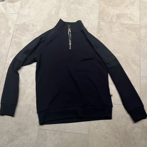 Svart tröja från Mos Mosh - Snygg svart tröja från Mos Mosh med dragkedja vid halsen och en stilren patch på ärmen. Perfekt för en avslappnad men ändå stilren look. KAN GÅ NER I PRIS VID SNABB AFFÄR