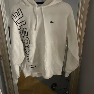 Vit hoodie från Lacoste - Säljer en snygg vit hoodie från Lacoste med stor logga på sidan och klassisk krokodil på bröstet. Hoodien har en känguruficka och justerbar huva med dragsko. Perfekt för en stilren och bekväm look.
