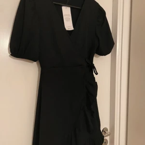 Svart omlottklänning - Elegant svart omlottklänning med korta ärmar och knytning i midjan. Klänningen har en v-ringad design och volangdetaljer längs kanten. Perfekt för en stilren look.