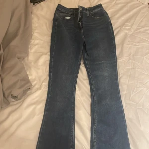 Blå bootcut jeans från Collusion - Snygga blå bootcut jeans från Collusion. De har en klassisk femficksdesign och en bekväm passform. Perfekta för en avslappnad stil med en touch av retro. Slitna detaljer vid fållen ger en cool look.