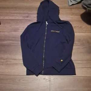 Mörkblå hoodie från Lyle & Scott - Säljer en snygg mörkblå hoodie från Lyle & Scott med dragkedja och huva. Tröjan har långa ärmar och en liten gul logga på ärmen. Perfekt för en avslappnad stil.