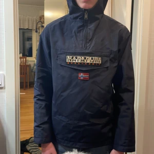 Svart vindjacka från Napapijri köpte den för 1300 säljer nu för 699 - Snygg svart vindjacka från Napapijri med en stor ficka framtill och dragkedja. Jackan har en huva och ett stilrent märke med flagga på bröstet. Perfekt för blåsiga dagar.