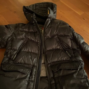 Svart dunjacka från Parajumpers - Säljer en svart dunjacka från Parajumpers med huva och flera praktiska fickor. Jackan har en quiltad design och dragkedja framtill. Perfekt för kyliga dagar. Den har ett hål (se sista bilden) som är lagat med laglapp för 3 månader sen därför har den rivits lite, men laglappar tillkommer med köp! Denna skada är ingenting som syns på avstånd endast om man söker efter den.