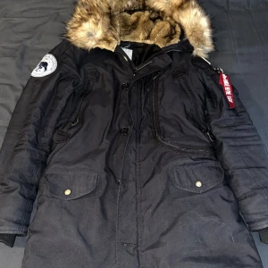 Svart parkajacka från Alpha Industries - Säljer en svart parkajacka från Alpha Industries med en mysig fuskpäls på huvan. Jackan har långa ärmar och flera praktiska fickor med knappar. Perfekt för kalla dagar med sin varma och skyddande design. Nypris 1800kr