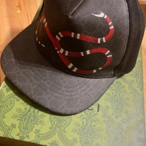 Svart keps med ormmönster - Snygg svart keps med ett coolt ormmönster i rött och vitt. Kepsen har en klassisk form med böjd skärm och mesh på baksidan för ventilation. Perfekt för att ge din outfit en edgy touch.