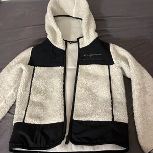 Vit fleecejacka med huva från Sail Racing - Säljer en vit fleecejacka från Sail Racing med svarta detaljer. Jag gillar värkligen denna jacka men den har blivit för liten för mig. Den är varm och skön och är väldig fin. Pris går att diskuteras. 