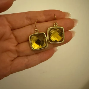 Guldigas örhängen med fyrkantig design - Eleganta örhängen i guld med fyrkantig form och facetterad yta. De har en krok för enkel användning och en glittrande gul detalj i mitten som fångar ljuset vackert.